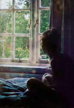 A girl sits crosslegged on a bed, looking out the window