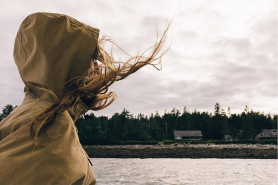 A woman in a hooded rain jacket looks towards a rocky Maine shore as her hair is blown by the wind